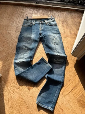 Blå slitna jeans med raka ben - Säljer ett par blå nudie jeans med slitningar och slim fit. Jeansen har klassisk femficksmodell, normal midja och ljus tvätt med distressed detaljer på framsidan. Perfekta för en avslappnad och trendig look.