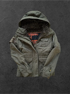 Fieldjacket Superdry xs passar S - Stilren grön vindjacka från Superdry med fyra framfickor, huva och mjukt foder i fejkpäls. Jackan har dragkedja, tryckknappar och detaljerade sömmar. Märkeslogga på bröstet och ärmen ger en cool touch. Perfekt för blåsiga dagar. Xs sitter som S