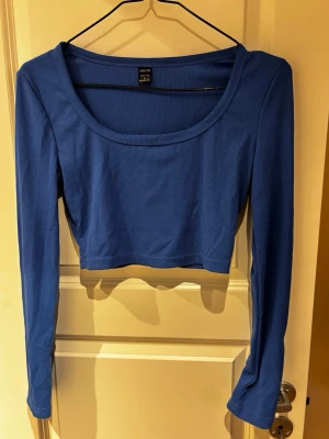 Blå ribbad croppad långärmad topp - Trendig blå croppad topp med lång ärm och rund halsringning. Tillverkad i ribbad bomullsmix som ger en skön och stretchig passform. Perfekt att styla med höga jeans eller kjol för en cool vardagslook.