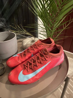 Nike Mercurial röda fotbollsskor - Säljer ett par Nike Mercurial fotbollsskor i rött med stora ljusblå swoosh-loggor på sidorna. Skorna har snörning och är tillverkade i ett lätt syntetmaterial som ger bra bollkontroll och passform. Perfekta för dig som vill ha speed och stil på planen.