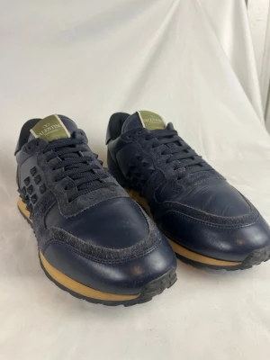 Valentino Garavani blå skor - Snygga mörkblå sneakers från Valentino Garavani med coola nitar på sidorna. Skorna har en mix av mocka och läder, svarta skosnören och beige sula. Perfekta för dig som vill sticka ut med en exklusiv och trendig look. Storlek 41 och i väldigt fint skick.