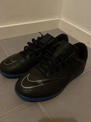 Svarta Nike fotbollsskor med blå sula - Säljer ett par svarta Nike futsalsskor med blå sula och stora Nike-loggor på sidan. Skorna har snörning och är tillverkade i syntetmaterial med en rund tå och platt sula. Insidan är blå och ovandelen har diskreta ränder för extra stil.