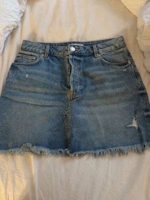 Blå jeanskjol med fransig kant - Säljer en blå jeanskjol med rå fransig kant och slitningar. Kjolen har klassisk femficksdesign, knapp och dragkedja framtill samt bälteshällor. Perfekt för en avslappnad och trendig look.