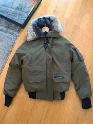 Grön pufferjacka från Canada Goose - Snygg grön pufferjacka från Canada Goose med stor huva och avtagbar fuskpäls. Jackan har svarta muddar, flera fickor och patch på ärmen. Perfekt för kalla vinterdagar och riktigt varm. Klassisk bombermodell med dragkedja och tryckknappar.