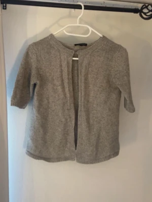 Grå kofta med kort ärm - Mysig grå kofta med korta ärmar och rund halsringning. Koftan är öppen och har en knapp högst upp. 