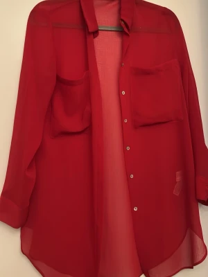 Röd transparent skjorta från Zara - Säljer en röd transparent skjorta från Zara med två stora bröstfickor och klassisk krage. Skjortan har knappar framtill och är tillverkad i ett lätt, strukturerat syntetmaterial. Perfekt för dig som vill ha en statement-piece i garderoben.
