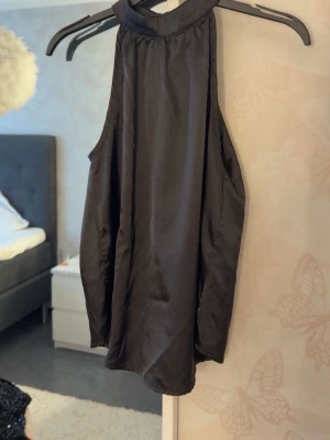 Svart ärmlös halterneck topp - Säljer en svart, ärmlös halterneck topp i glansigt material. Toppen har hög krage och ett elegant, löst fall. Perfekt att styla med jeans eller kjol för en trendig look.