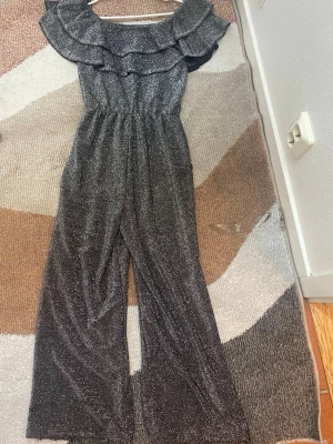 Glittrig svart jumpsuit med volang - Snygg svart jumpsuit med glittereffekt och breda ben. Off shoulder-modell med dubbla volanger upptill som ger en trendig look. Resår i midjan för skön passform. Perfekt för dig som vill sticka ut lite extra. Storleken 152 men den passar mig trots att jag har växt (är 157)