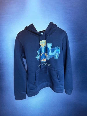 Mörkblå hoodie Polo Ralph Lauren - Mörkblå hoodie från Polo Ralph Lauren med ikoniskt björnmotiv och bil på bröstet. Klassisk känguruficka framtill, justerbar huva med snören och mjukt material. Perfekt för en avslappnad och trendig stil.