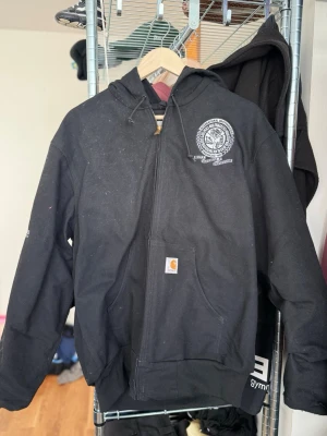 Vintage Carhartt jacka - Svart Carhartt zip jacka i storlek M (herr). Tjock och slitstark med klassisk logga, perfekt för en clean streetwear-look.