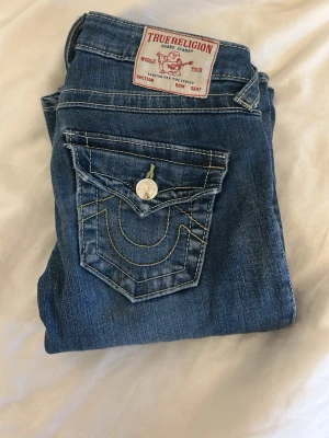 Blå jeans True Religion med fickdetalj - Säljer ett par blå jeans från True Religion med ikonisk fickdetalj och kontrastsömmar. Klassisk femficksmodell med silverfärgade knappar och patch bak i midjan. Jeansen har en snygg tvätt och raka ben. Perfekta för dig som gillar street och premium denim.
