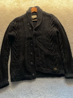 Svart stickad Ralph Lauren kofta med mönster - Säljer en svart stickad kofta med grovt strukturmönster och sjalkrage. Koftan har knäppning framtill med fem knappar, två fickor och ribbade muddar. Perfekt för dig som gillar en klassisk men ändå unik look. Storleken är s men passar även mig som en  medium. Hör av er vid minsta fundering mvh Artur !