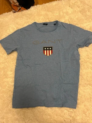 Blå GANT t-shirt med logga - Snygg blå t-shirt från GANT med klassisk rund hals och korta ärmar. Framtill finns ett tryck med GANT-loggan och ett sköldmärke med stjärnor och ränder i rött, vitt och blått. Perfekt till jeans eller shorts för en avslappnad look.
