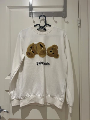 Vit Palm Angels sweatshirt med björn - Säljer en vit sweatshirt från Palm Angels med det ikoniska björnmotivet i beige och svart brodyr på bröstet. Tröjan har rund halsringning och ribbade muddar vid ärmar och nederkant. Perfekt statement-plagg för dig som gillar streetwear och coola detaljer.