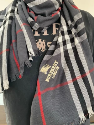 Grå rutig halsduk från Burberryy - Snygg grå halsduk från Burberryy med klassiskt rutmönster i svart, vitt och rött. Halsduken har fransade kanter och broderad Burberryy London-logga i gult. Perfekt accessoar för att lyfta din outfit.