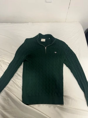 Mörkgrön kabelstickad tröja Gant - Snygg mörkgrön kabelstickad tröja från Gant med halv dragkedja och liten broderad logga på bröstet. Tröjan har ribbade muddar och hög krage, perfekt för lager på lager. Klassisk och stilren design som funkar till det mesta.