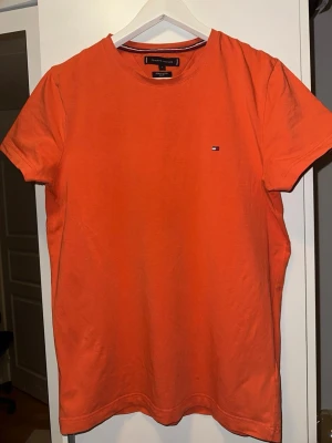 Orange t-shirt från Tommy Hilfiger - Snygg ljus orange t-shirt från Tommy Hilfiger i ekologisk bomull. Slim fit-modell med korta ärmar och rund hals. Liten klassisk Hilfiger-logga broderad på bröstet. Perfekt för en clean och stilren look.