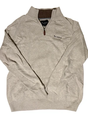 Beige halvzip tröja från Bondelid - Stilren beige stickad tröja från Bondelid med halvzip och hög krage. Tröjan har diskret broderad logga på bröstet och ribbade muddar vid ärmslut och nederkant. Perfekt för lager-på-lager och enkel att matcha med olika outfits.