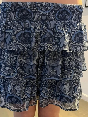 Blå volangkjol med mönster - Kort kjol med flera volanger i blått och vitt mönster. Kjolen har bred resår i midjan och ett lätt, luftigt tyg som ger en flowy känsla. Perfekt för dig som gillar en feminin och trendig stil.