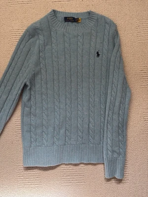 Ljusblå kabelstickad tröja Polo Ralph Lauren - Kabelstickad tröja i ljusblått från Polo Ralph Lauren  med broderad marinblå logga på bröstet. Klassisk och stilren design i mjukt material.