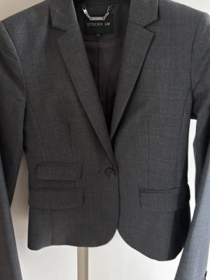 Mörkgrå kavaj från STOCKH LM - Snygg mörkgrå kavaj från STOCKH LM med klassisk krage, enkelknäppning och två fickor framtill. Kavajen har en figurnära passform och är helfodrad med slät insida. Perfekt för en stilren look.