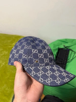 Blå GG-mönstrad keps från Gucci - Snygg blå keps från Gucci med klassiskt GG-mönster över hela kepsen. Tillverkad i denimliknande material med justerbar rem bak i blått läder och gulddetalj. Perfekt för dig som vill sticka ut med en lyxig accessoar.