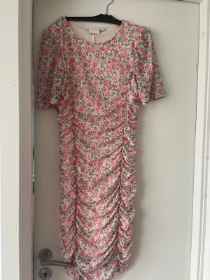 Rosa blommig midiklänning från Vila  - Supersöt midiklänning från Vila  med rosa och grönt blommönster på vit botten. Klänningen har rynkade detaljer framtill och baktill, rund halsringning och korta ärmar. Perfekt för dig som gillar romantisk och trendig stil. Nästan oanvänd, har endast hafts på ett fåtal gånger. 