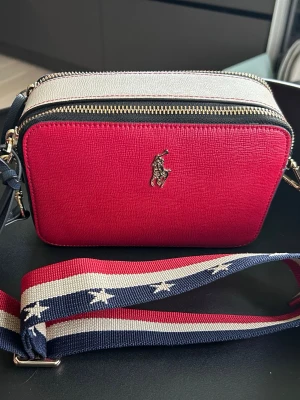 Röd axelväska från Polo Ralph Lauren - Snygg röd axelväska från Polo Ralph Lauren. Väskan har två fack och ett mindre med dragkedja inuti samt en bred axelrem framtill, två dragkedjor och en bred axelrem. Perfekt storlek (29x13 cm)för det viktigaste och ger en cool touch till din outfit. Använd vid ett tillfälle, som ny.