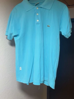 Ljusblå pikétröja från Lacoste - Klassisk ljusblå pikétröja från Lacoste med korta ärmar, krage och knappar framtill. Tröjan har den ikoniska krokodilloggan på bröstet och är tillverkad i mjuk bomull. Perfekt för en avslappnad och stilren look.