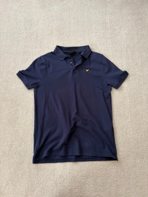 Marinblå pikétröja från Lyle & Scott - Snygg marinblå pikétröja från Lyle & Scott med klassisk krage och knappar framtill. Tröjan har det ikoniska gula fågelmärket broderat på bröstet och är tillverkad i mjuk bomull. Perfekt till jeans eller shorts för en clean look.