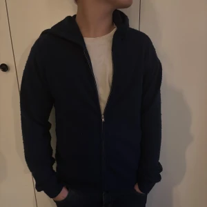 Mörkblå kashmir hoodie från RYVLS med dragkedja - Mysig mörkblå hoodie från RYVLS med hel dragkedja framtill och huva. Tröjan är tillverkad i ett mjukt, stickat material och har långa ärmar. Perfekt för kyliga dagar och enkel att matcha med jeans eller joggers. Kashmir Grisch Laidback! Stor i storlek, han i bilden är 164 och sitter fint. Passar dels folk från 155-170