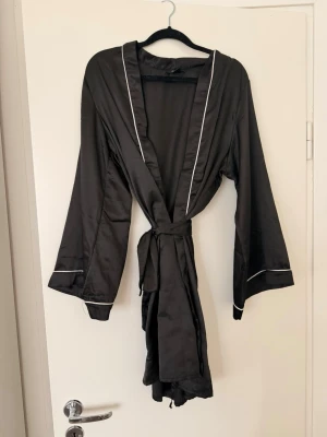 Svart satin morgonrock med vita detaljer - Stilren svart morgonrock i glansigt satinmaterial med vita passpoaldetaljer längs krage och ärmslut. Knytskärp i midjan och långa ärmar ger en avslappnad och lyxig känsla. Perfekt för mysiga hemmakvällar. Står storlek L men skulle säga mer S.