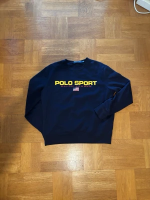 Mörkblå sweatshirt Polo Sport - Mörkblå sweatshirt från Polo Sport Ralph Lauren med gul logotyp och amerikansk flagga på bröstet. Tröjan har rund halsringning och långa ärmar. Köpte på outlet i New York i somras. 