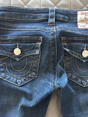 True Religion blå jeans med kontrastsömmar - Snygga blå jeans från True Religion med ikoniska kontrastsömmar och detaljerade bakfickor med lock och silverfärgade knappar. Klassisk femficksmodell i denim med raka ben och låg midja. Köpte för 1300kr 