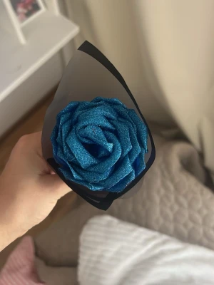 Blå glittrig konstgjord ros - Säljer en konstgjord ros i starkt blå färg med glittereffekt. Rosen är inslagen i svart och transparent papper och passar perfekt som dekoration eller present. Materialet är troligtvis plast eller skum med glittrig yta.