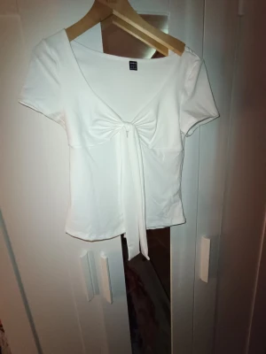 Vit topp med knyt framtill - Snygg vit topp med korta ärmar och knyt framtill som ger en trendig look. Toppen har en djupare ringning och är tillverkad i ett mjukt, stretchigt material som sitter bekvämt. Perfekt att styla med jeans eller kjol för en fräsch outfit.