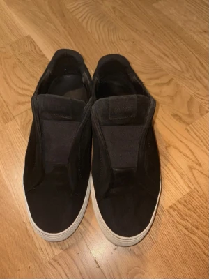 Svarta sneakers från Axel Arigato - Snygga svarta slip-on sneakers från Axel Arigato i mocka med vit sula. Diskret logga i guld på sidan och elastiska paneler för enkel påtagning. Perfekta för en clean och stilren look.