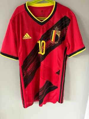 Belgien fotbollströja Hazard #10 Adidas - Röd Belgien landslagströja från Adidas med svarta och gula detaljer, nummer 10 och E. Hazard på ryggen. Tröjan har V-ringning, korta ärmar och klubbmärke på bröstet. Perfekt för fotbollsfans som vill sticka ut på matchen.
