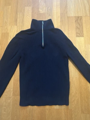 Mörkblå halvzip tröja East West - Mörkblå stickad tröja från East West med hög krage och silverfärgad halvzip. Tröjan har lång ärm och ribbstickad struktur, vilket ger en clean och stilren look. Perfekt för lager-på-lager och funkar till det mesta.