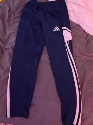 Mörkblå Adidas mjukisbyxor/ träningsbyxor  - Säljer ett par mörkblå mjukisbyxor från Adidas med vita detaljer och klassiska ränder längs benen. Byxorna har resår i midjan och är tillverkade i ett mjukt och bekvämt material, perfekta för chill dagar eller träning. Adidas-logga på låret för en sportig look. PRISET KAN DISKUTERAS!