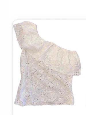 Vit oneshoulder blus med broderi - Superfin vit blus med oneshoulder och volangdetalj. Blusen har broderade blommor och är perfekt för dig som gillar romantisk stil. Luftigt material som passar bra till både jeans och kjol.