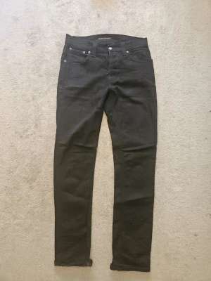 Svarta jeans från Nudie Jeans - Snygga svarta jeans från Nudie Jeans med klassisk femficksdesign och raka ben. Jeansen har silverfärgade nitar och knapp, samt diskret logga på bakfickan. Perfekta för en stilren och enkel look.