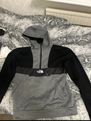 Grå och svart hoodie från The North Face - Snygg hoodie från The North Face i grått och svart med halv dragkedja och huva. Logotypen är broderad på bröstet. Materialet är mjukt och bekvämt, perfekt för chill eller streetwear. Stora fickor framtill och cool design med kontrastpaneler.