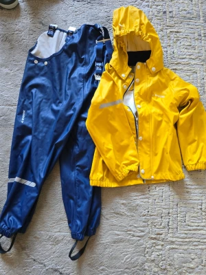 Gul regnjacka från Everest - Gul regnjacka från Everest med huva, reflexdetaljer och dragkedja med tryckknappar. Jackan har elastiska muddar vid ärmslut och nederkant samt vattenavvisande material. Perfekt för regniga dagar. Storlek 110/116 