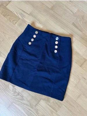 Mörkblå kjol med silvriga knappar - Snygg mörkblå kortkjol med dekorativa silvriga knappar framtill i två rader. Kjolen har en rak passform och är perfekt för dig som gillar en stilren look. Materialet känns som bomull eller polyester och kjolen har en klassisk midjehöjd.