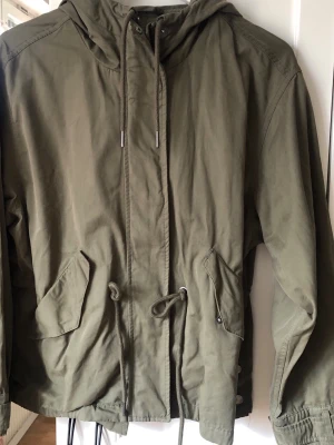 Militärgrön parkas med huva och dragsko - Snygg militärgrön parkas med huva och dragsko i midjan. Jackan har två stora fickor framtill, knappar och dragkedja samt justerbar nederkant. Perfekt för lager på lager och streetstyle. Materialet känns slitstarkt och jackan har en avslappnad passform. Fler fina detaljer, se bilder.