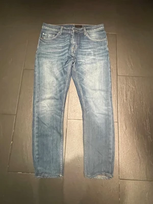 Blå jeans med fade - Säljer ett par blå jeans med klassisk femficksdesign och snygga slitningar på framsidan. Jeansen har raka ben och normal midja. Materialet är denim och färgen är ljusblå med tvättade detaljer. Perfekt för dig som gillar en avslappnad och trendig look. Storlekslappen är avklippt men passar runt 175-185 gissar på 30/31