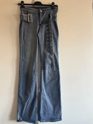 Blå bootcut jeans med bälte - Säljer ett par blå bootcut jeans med hög midja och tillhörande denimskärp med metallspänne. Jeansen har klassiska fem fickor och knappgylf. Materialet är denim i 100% bomull och tvätten är ljusblå med lätt slitning. Perfekta för en trendig och avslappnad look.