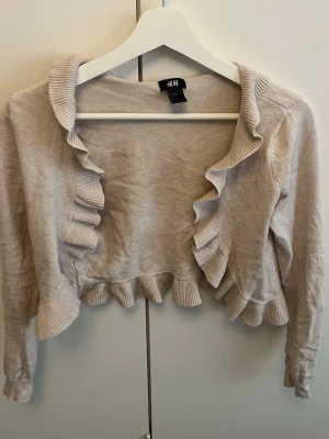 Beige volangkofta från H&M - Supersöt beige kofta från H&M med volangdetaljer längs krage, framkant och nederkant. Kort modell med långa ärmar, perfekt att slänga över en topp för en extra touch. Mjuk och lättstickad känsla.