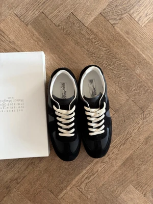Svarta Maison Margielas  - Snygga svarta sneakers från Maison Margiela med vita kontrasterande skosnören och gummisula i beige. Ovandelen är i en mix av mocka och läder, vilket ger en stilren och exklusiv look. Klassisk låg modell som passar till många outfits.